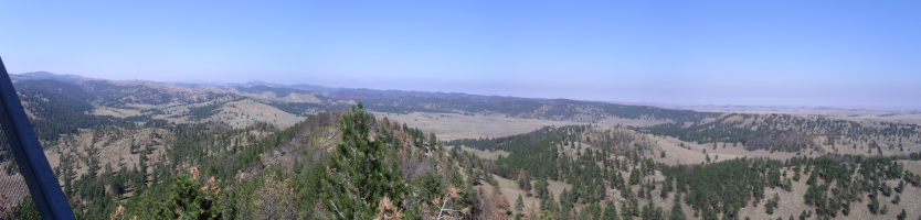 [About a dozen photos stitched together in three parts. This first part starts with the outer edge of the railing on the left and has an expanse of many mountains with trees on them providing splotches of green color on an otherwise light  tan ground. ]